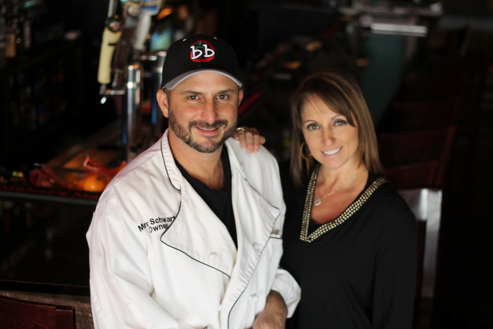 A man in chef’s whites and a cap standing with a woman in black inside the restaurant