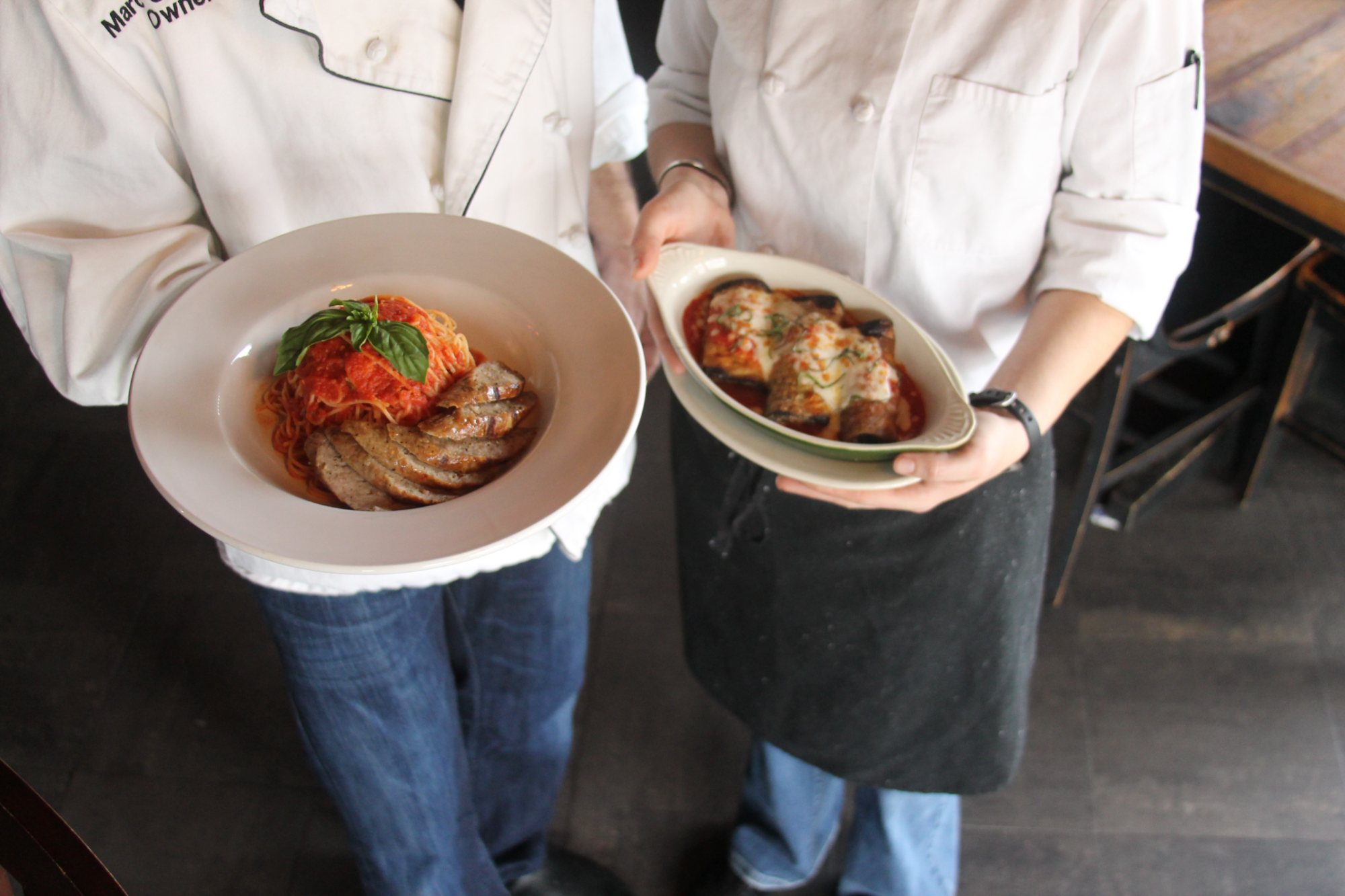 Two servers carrying shallow bowls of pasta, one topped with red sauce and basil