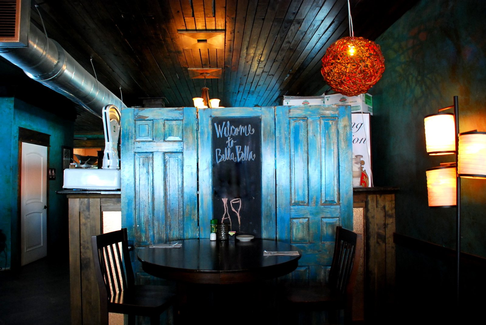 A table set in front of painted blue doors, beside a chalkboard welcome sign and a round orange lamp
